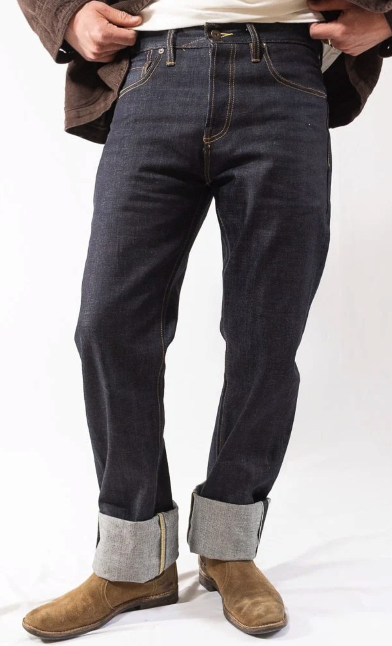 Man wearing dark indigo raw selvedge straight leg jeans with gold stitching, cuffed at the hem, paired with suede boots and a brown jacket