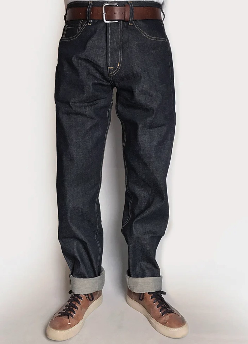 Man wearing dark indigo relaxed fit raw denim jeans with white stitching, brown leather belt, and cuffed selvedge hem, paired with brown sneakers